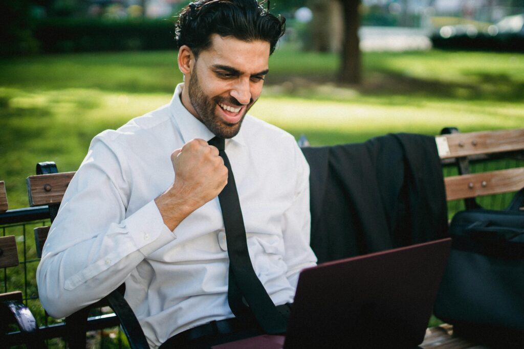 A joyful businessman celebrates a success working on his laptop outdoors on a park bench.